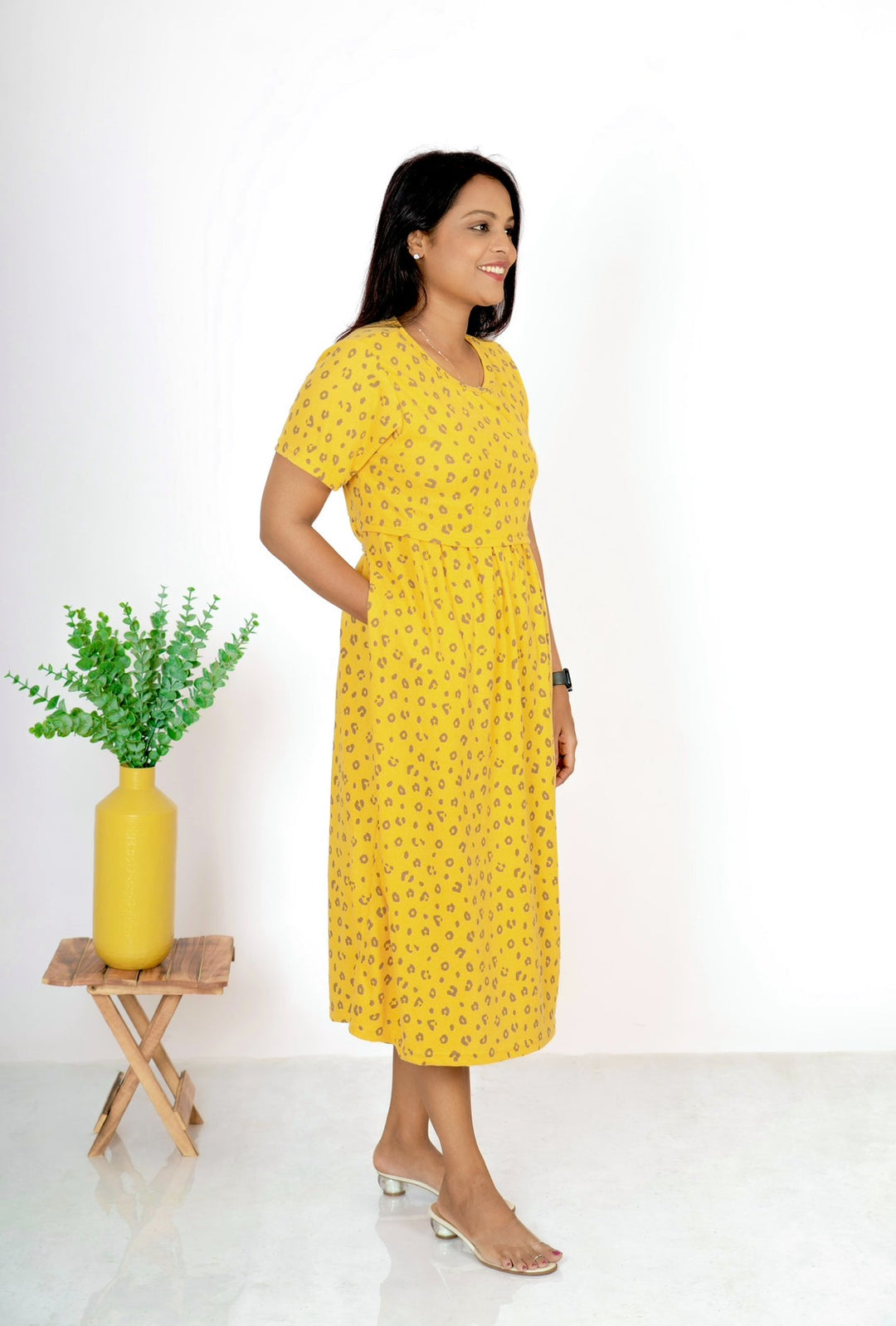 Woman wearing ultra soft midi yellow waffle nursing dress with flower print and round neck standing indoors next to a small wooden table with a yellow vase and green plant