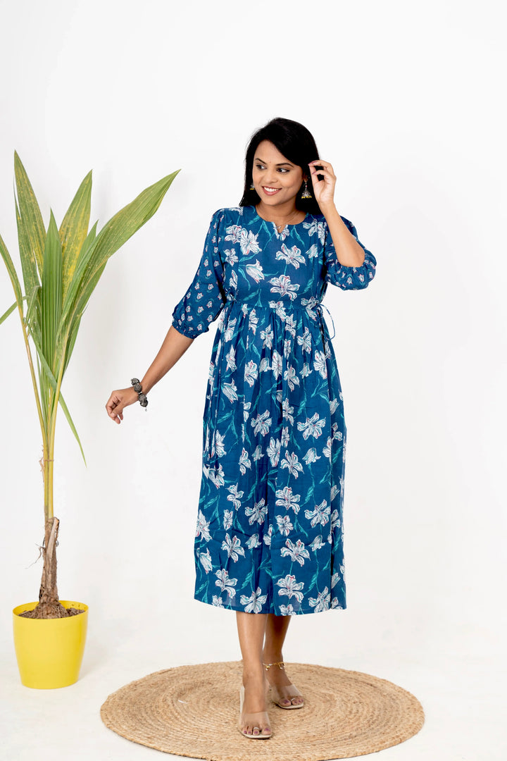 Woman wearing blue floral Nivi day to night cotton dress with three-quarter sleeves and round neckline, standing indoors near a plant.