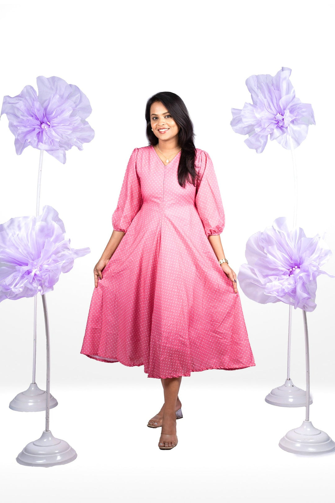 Woman wearing pink cotton Candy casual dress with V-neck, 3/4 sleeves, dot print, and center zipper, standing between purple floral decorations.