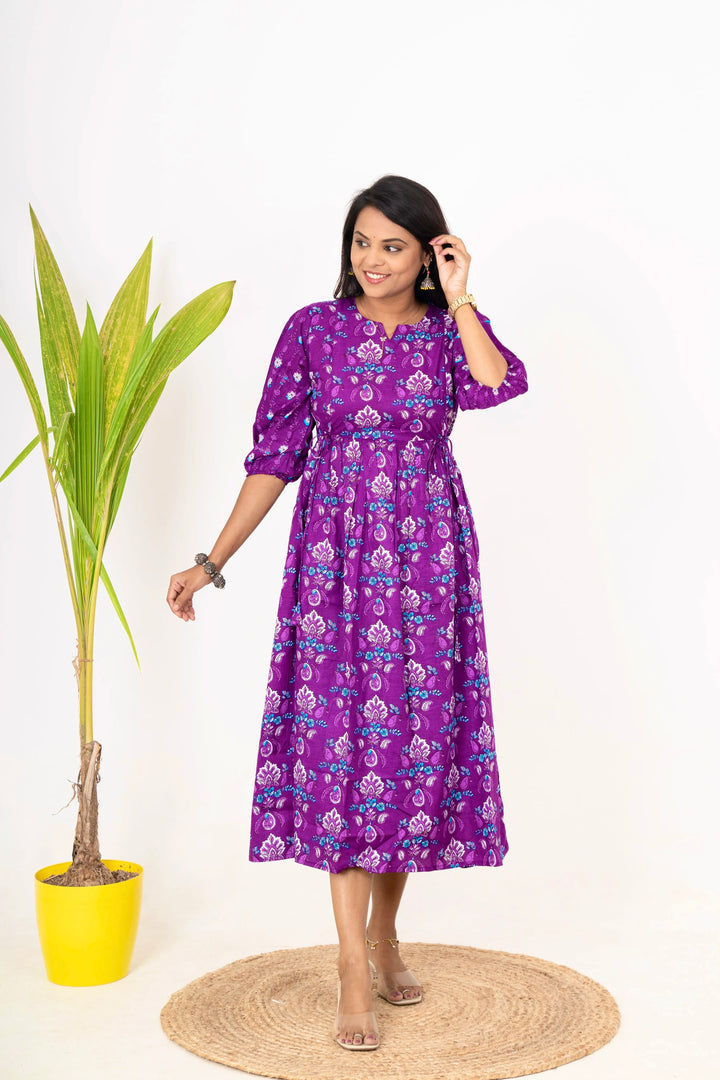 Woman wearing purple floral Janani maternity day to night dress with 3/4 sleeves and round neckline standing beside a potted plant