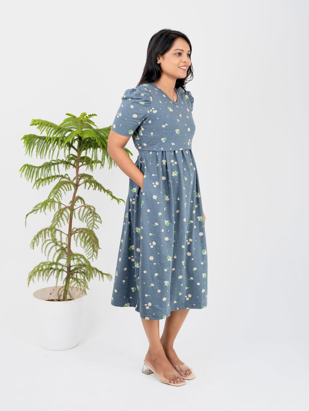 Woman wearing a blue floral Midi Ridha Light Weight nursing dress with elbow-length puffy sleeves and V-neck, standing beside a potted plant.