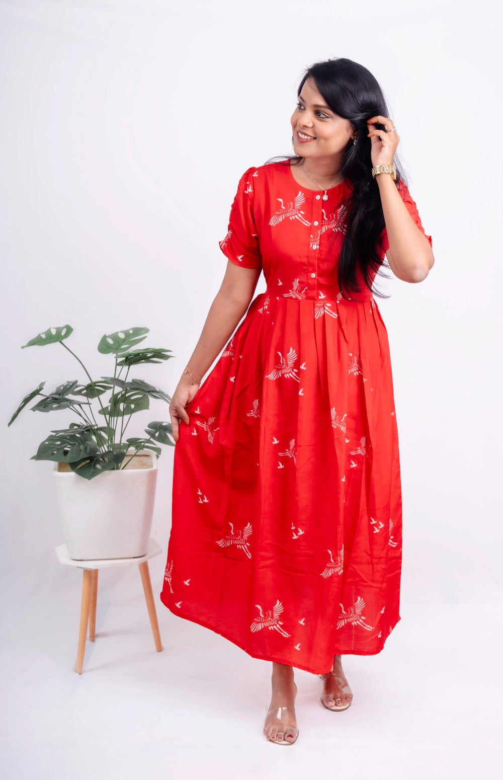 Woman wearing red day to night dress with floral print and fish cut sleeves standing next to a potted plant