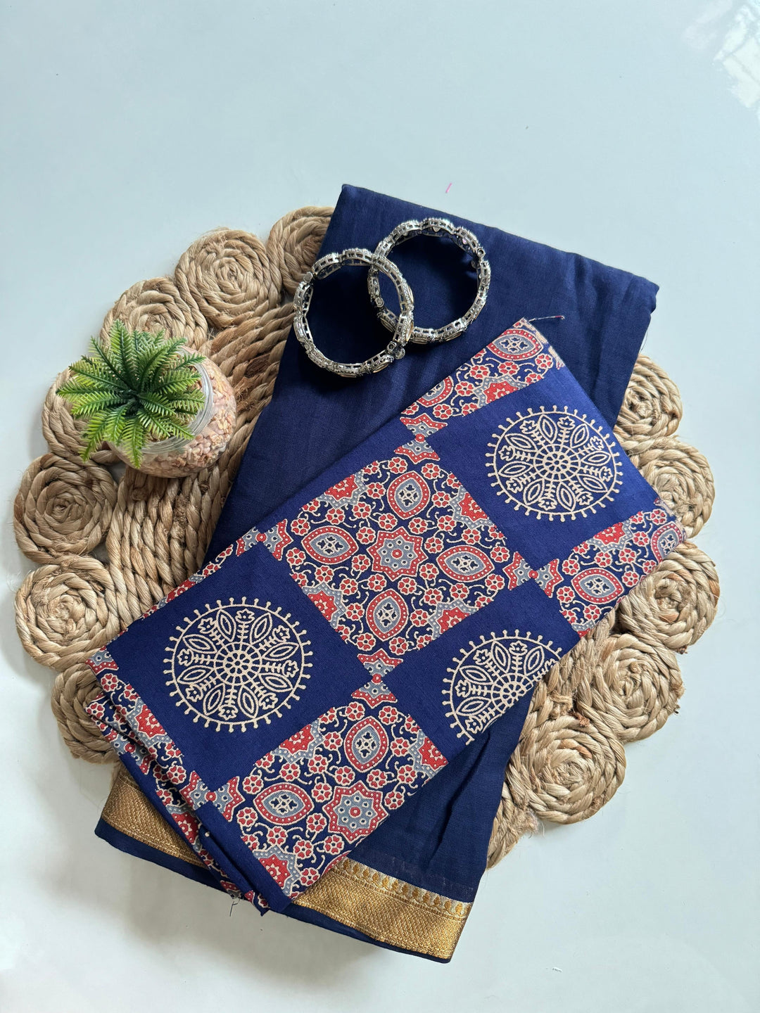 Blue mul cotton saree with intricate beige and orange block prints and gold border, paired with silver bangles on woven mat with small green succulent plant