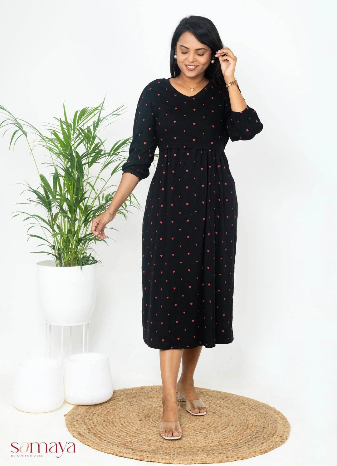 Woman wearing black midi ribbed dress with red heart print and 3/4 sleeves standing indoors next to a potted plant