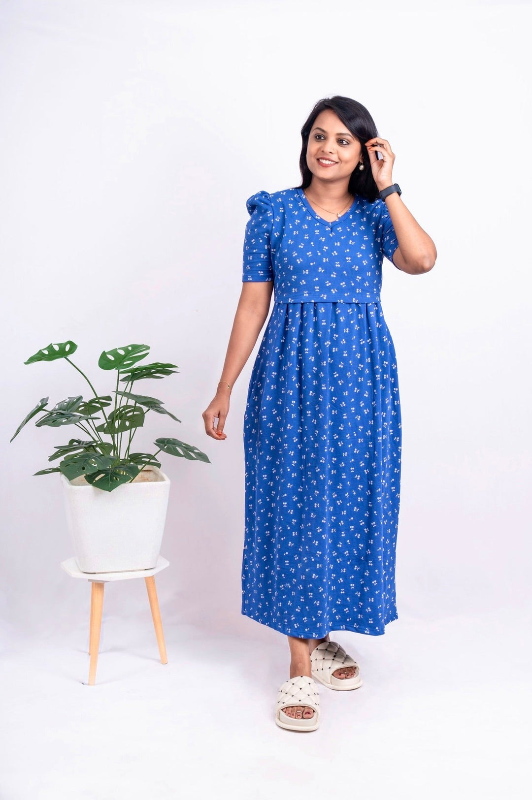 Woman wearing a blue cherry floral print maxi nursing dress with puff elbow sleeves and V-neckline standing next to a potted plant