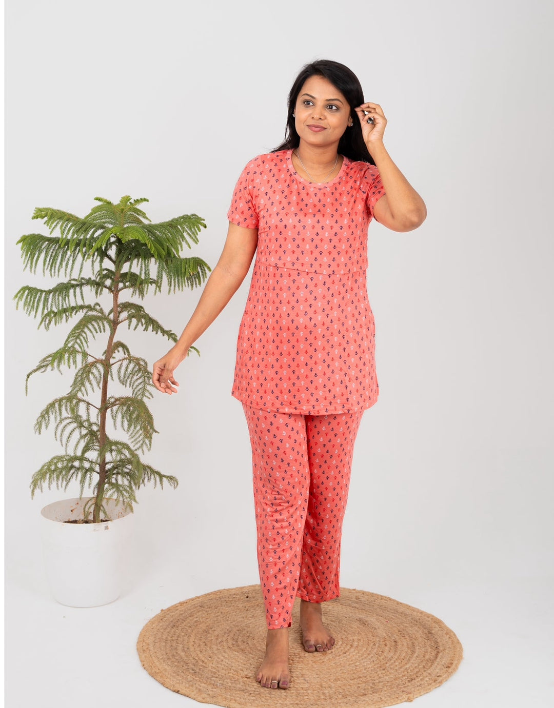 Woman wearing orange floral nursing top and pant set with short sleeves and round neck, standing barefoot on a circular woven mat next to a potted green plant