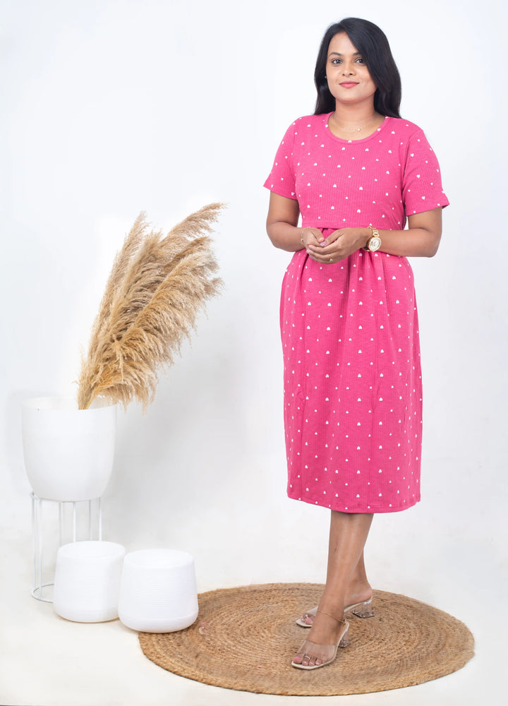 Woman wearing pink Midi Rosey Ribbed Dress with white heart print, short sleeves, and round neck, standing indoors.