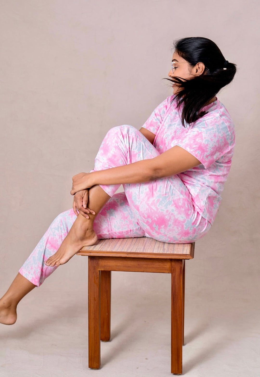 Woman wearing pink patterned candy feeding pant set sitting sideways on wooden stool against plain background