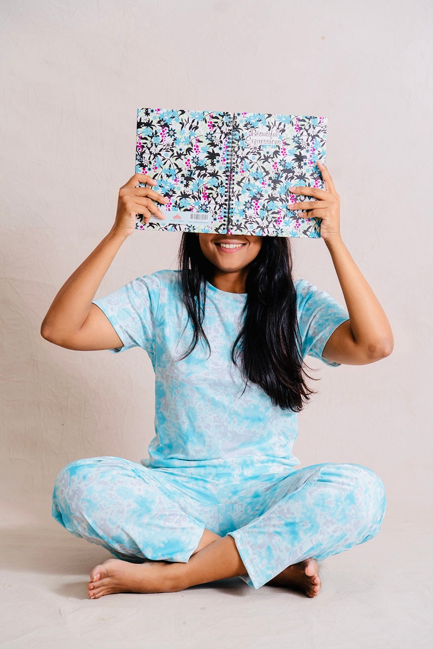 Woman wearing relaxed fit aqua pant set sitting cross-legged holding a floral notebook above her face